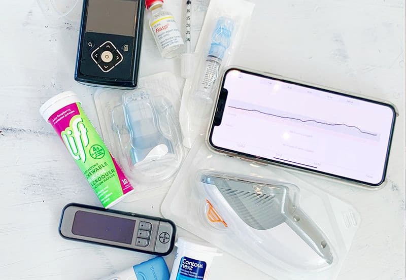 Una bomba de insulina, un iPhone con gráficos de CGM, jeringas, viales de insulina y un medidor de glucosa sobre la mesa.