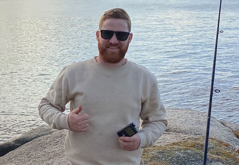 Andrew Burgess ina Sheasamh ar Charraig Cois Uisce ag Taispeáint a Chaidéil Inslin